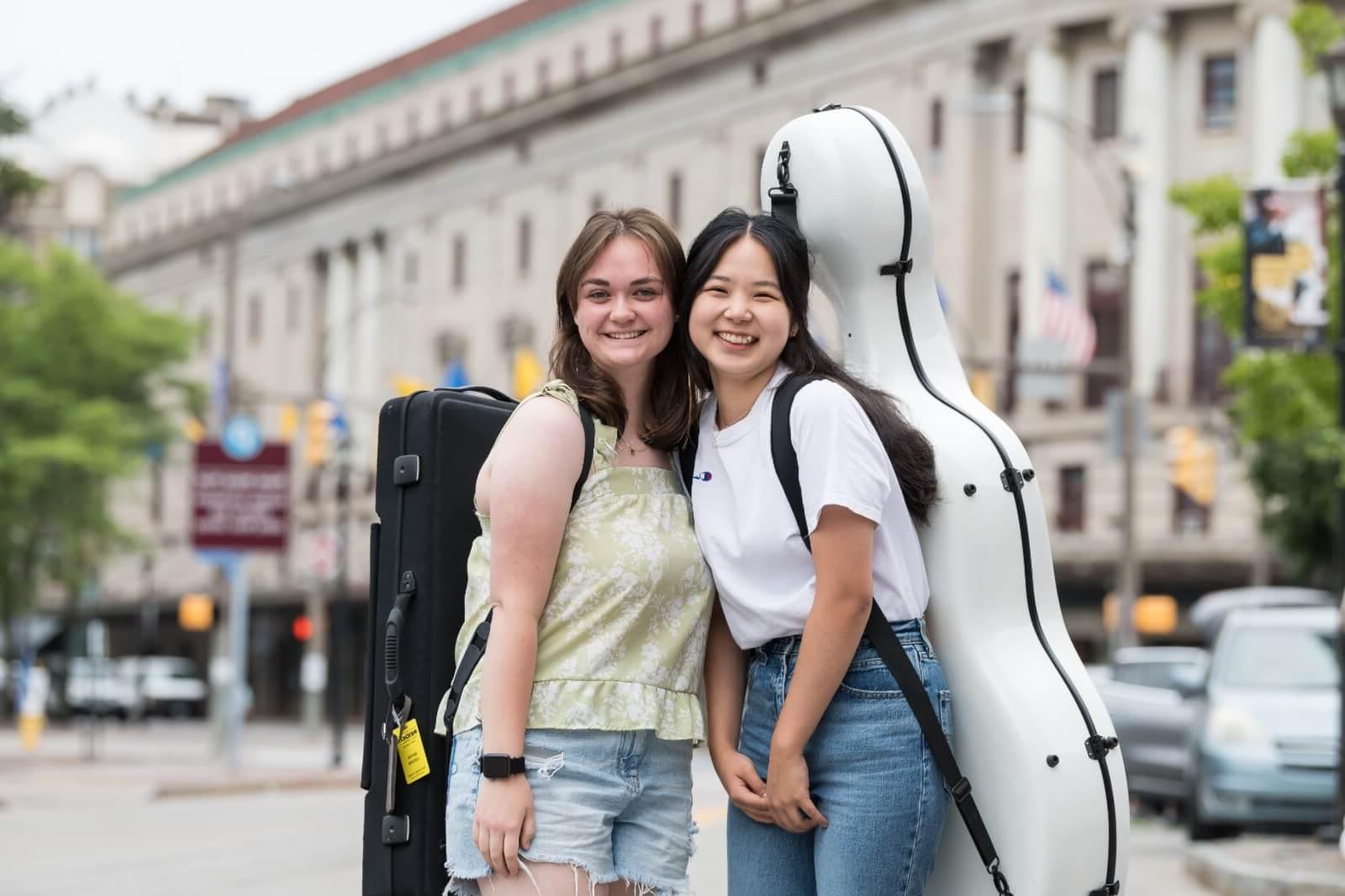 Two smiling young women stand close together, each carrying instrument cases on their backs. They are outdoors in an urban area with blurred buildings, vehicles, and street signs in the background. One wears a white shirt and jeans, the other a green top and shorts.