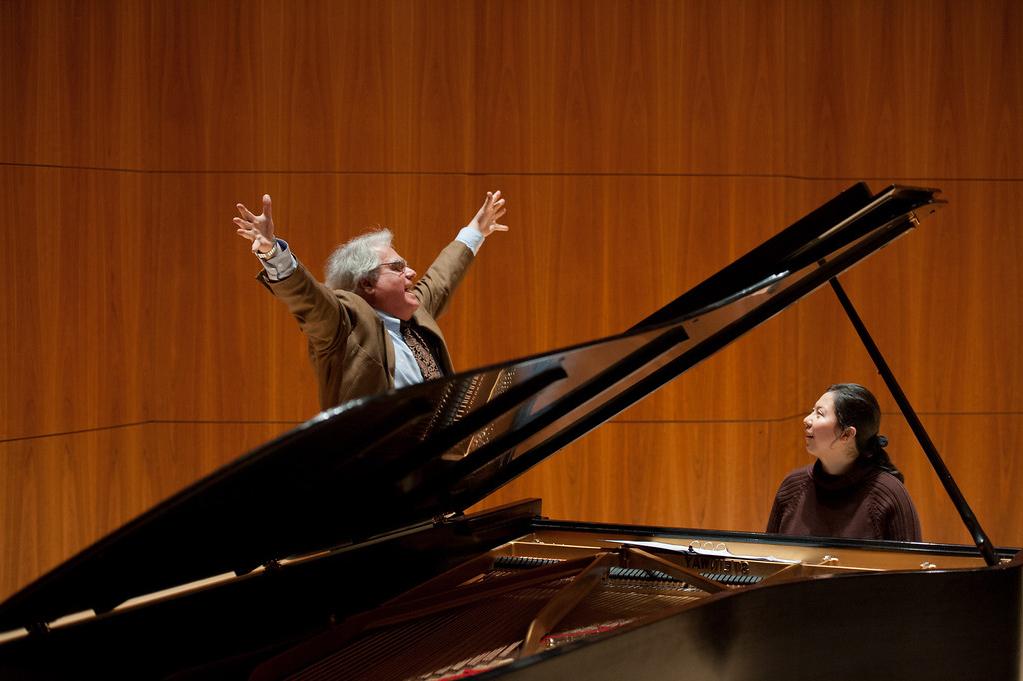 A musician stands triumphantly over a piano with his arms spread out, with another person sitting playing the piano